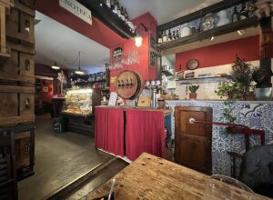 Inside a red-walled Florence wine bar with an enoteca sign, a barrel-fronted tap, bottles on shelves, a glass cabinet and tiled counter.