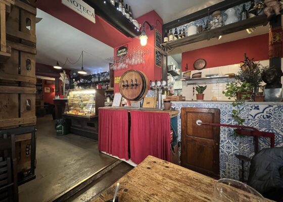 Inside a red-walled Florence wine bar with an enoteca sign, a barrel-fronted tap, bottles on shelves, a glass cabinet and tiled counter.
