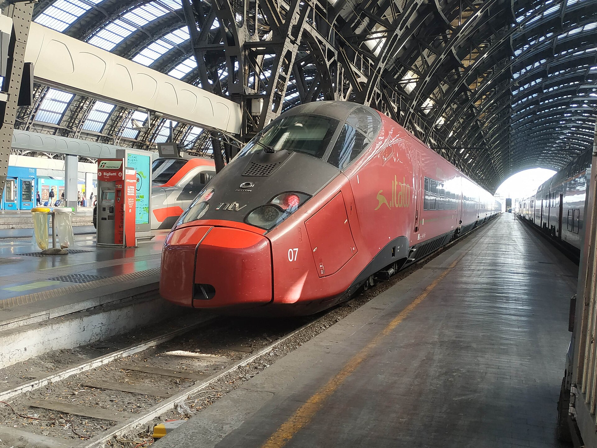 A red Italo high-speed train with a long pointed nose at a platform under the arched roof of Milano Centrale