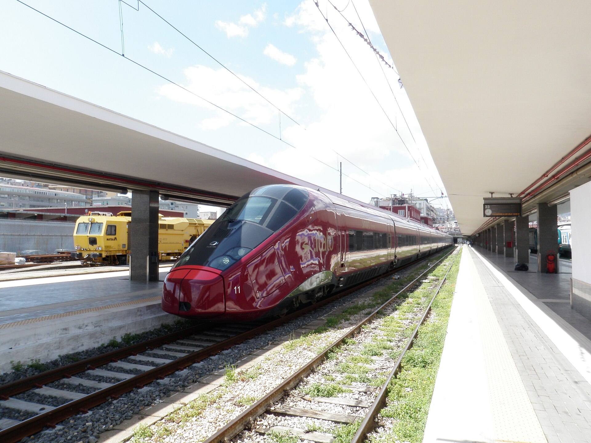 A dark red Italo high-speed train with a sloping nose standing at a platform in Naples, with yellow maintenance wagons on the next track