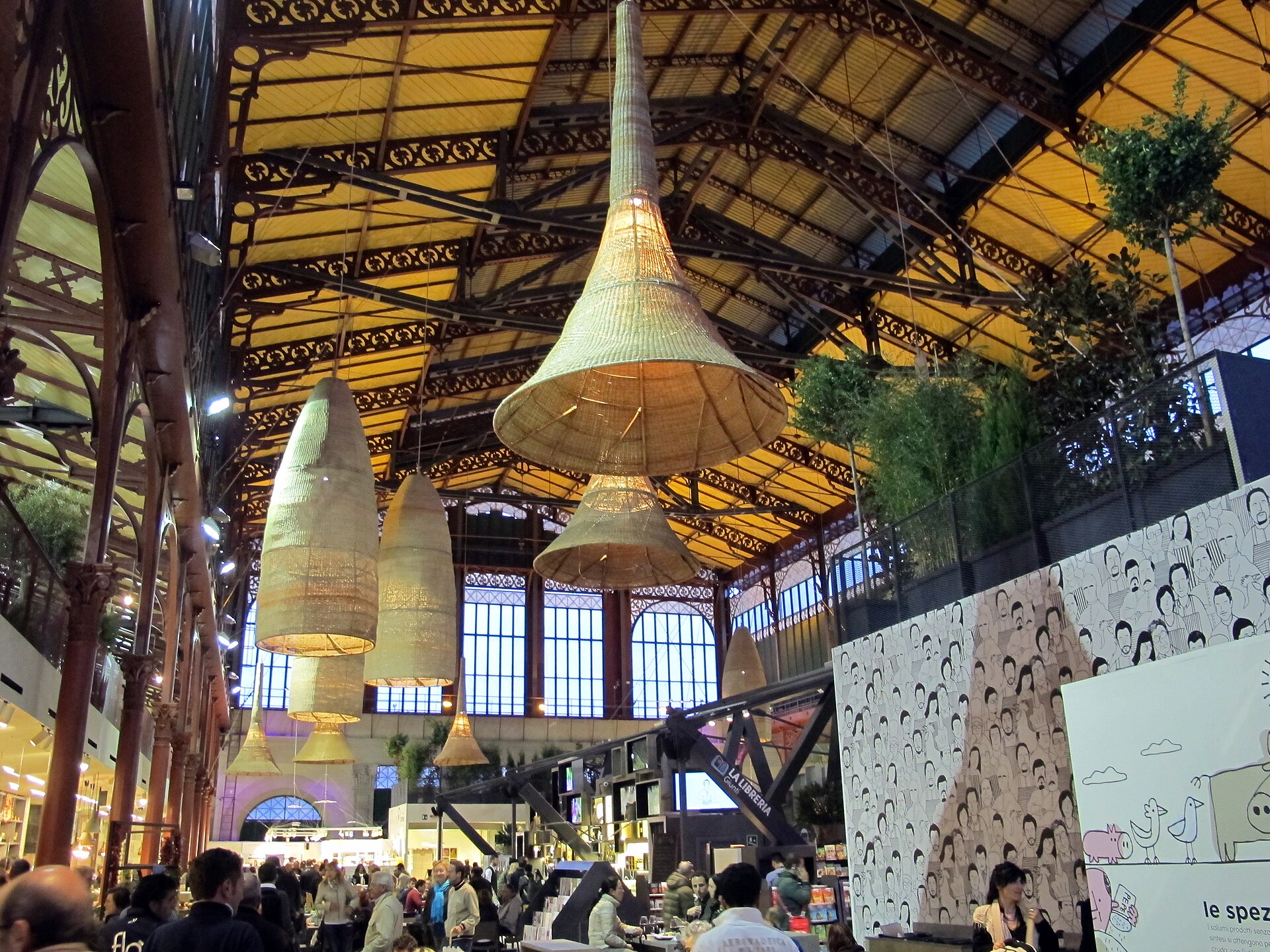 Large hanging lamps illuminate visitors and food counters beneath the iron roof of Florence's Mercato Centrale upstairs food hall.