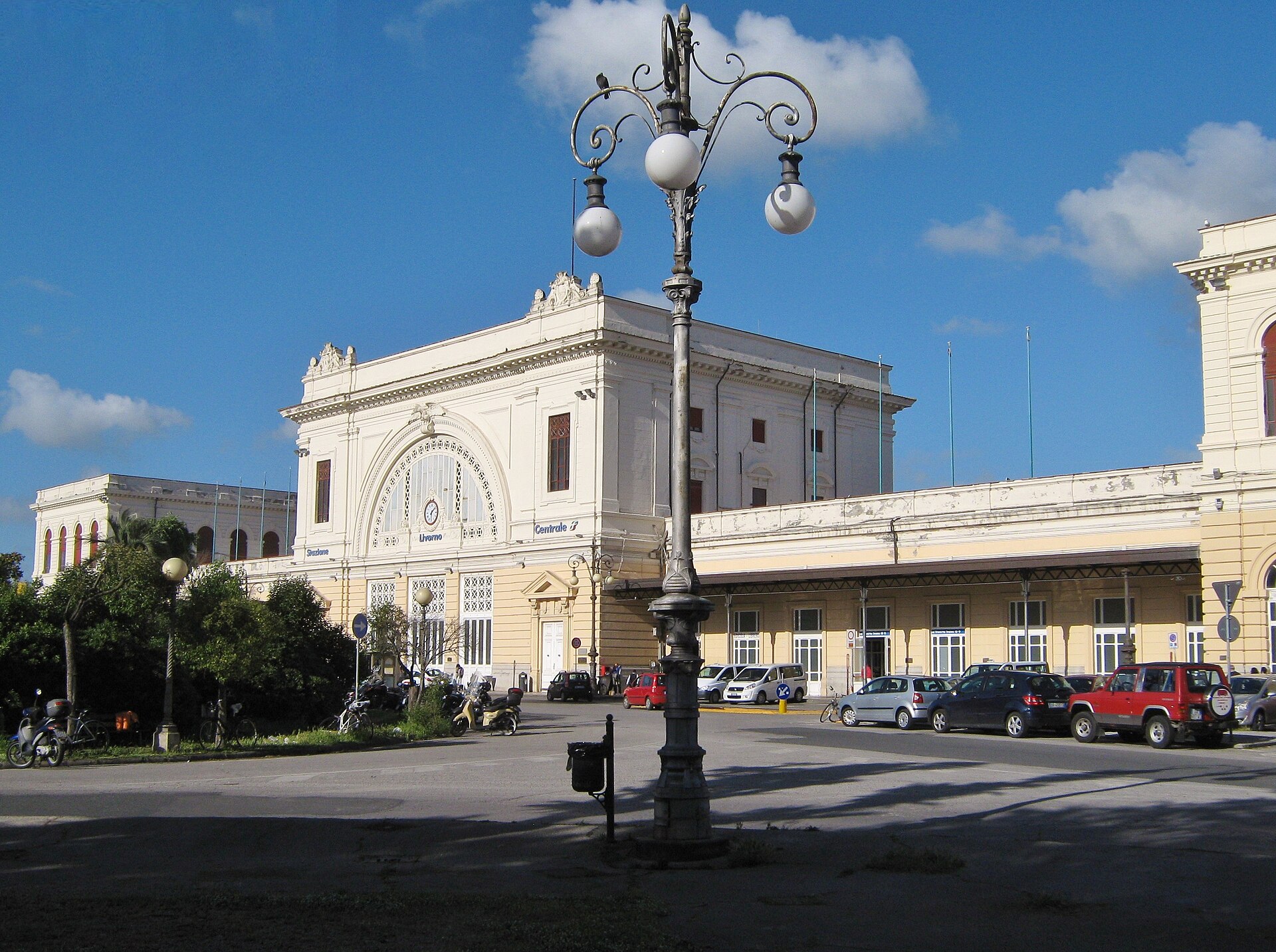 The cream neoclassical facade of Livorno Centrale railway station behind an ornate lamp post, with parked cars in the forecourt under a blue sky