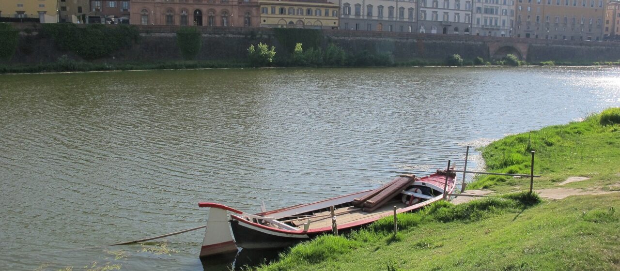 A long wooden barchetto with red trim lies moored beside a grassy bank of the Arno in Florence.