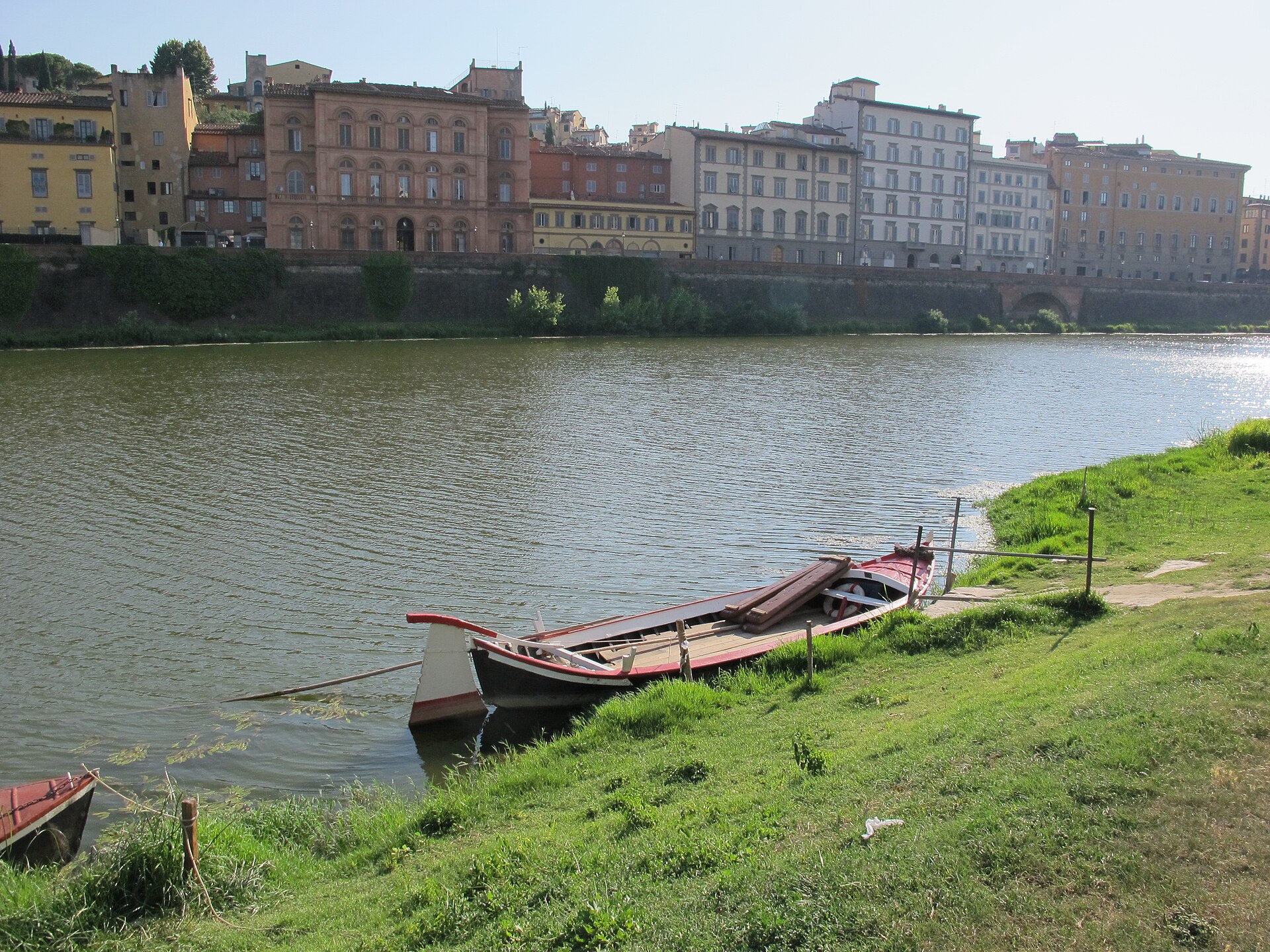 A long wooden barchetto with red trim lies moored beside a grassy bank of the Arno in Florence.