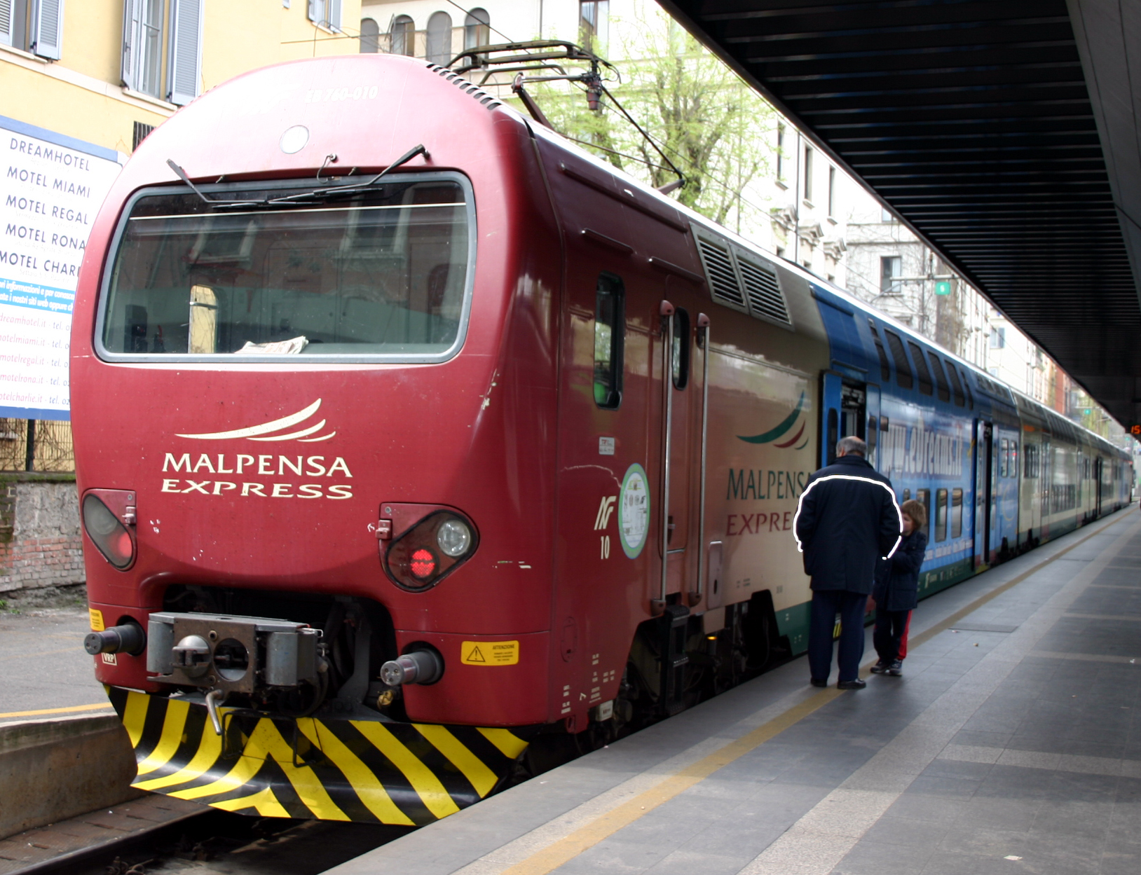 A red Malpensa Express train with a white logo on its cab, standing at a platform with a man in a dark jacket beside it