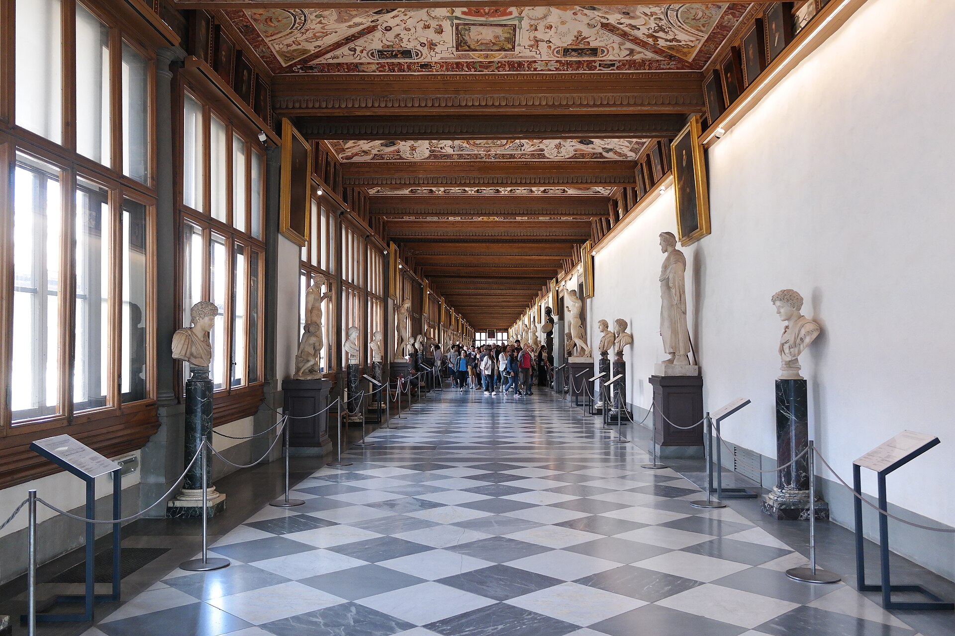 Marble statues and busts line the Uffizi's east corridor in Florence beneath a painted ceiling, with visitors in the distance.