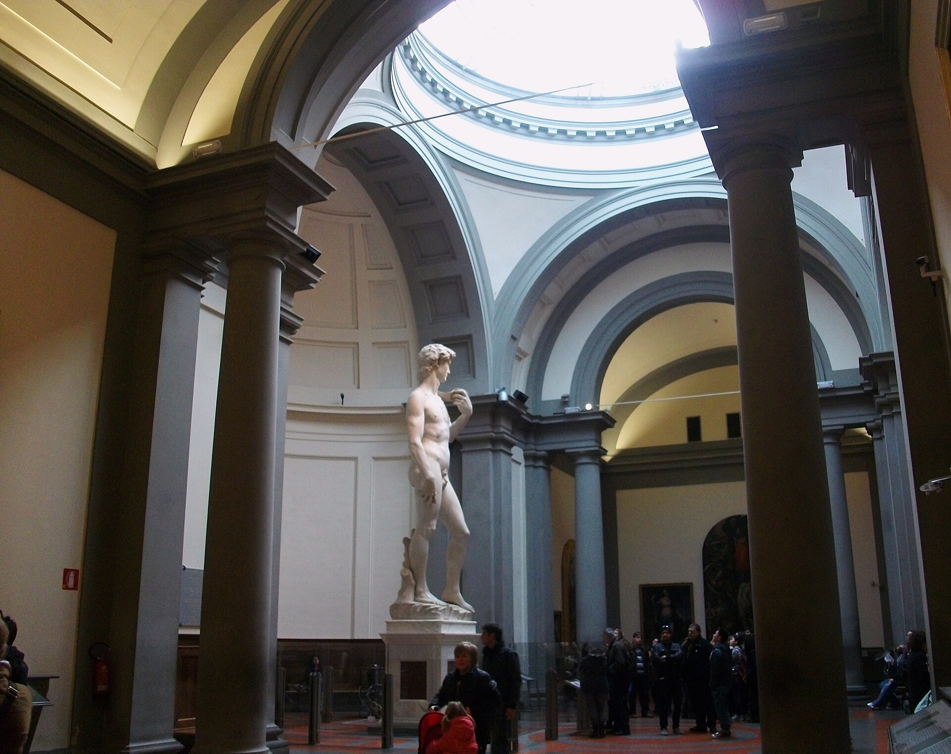 Michelangelo's David stands beneath the circular skylight in Florence's Accademia, framed by columns with visitors below.