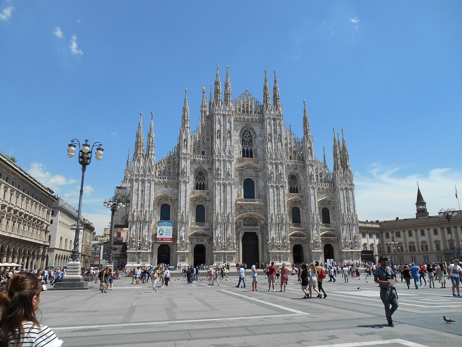 The white marble Gothic facade of Milan cathedral with its many spires, seen across the busy Piazza del Duomo on a sunny day