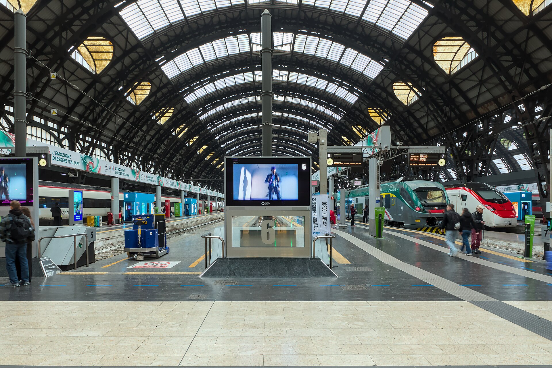 The platform concourse of Milano Centrale under its iron and glass roof, with information screens and trains waiting at the buffers