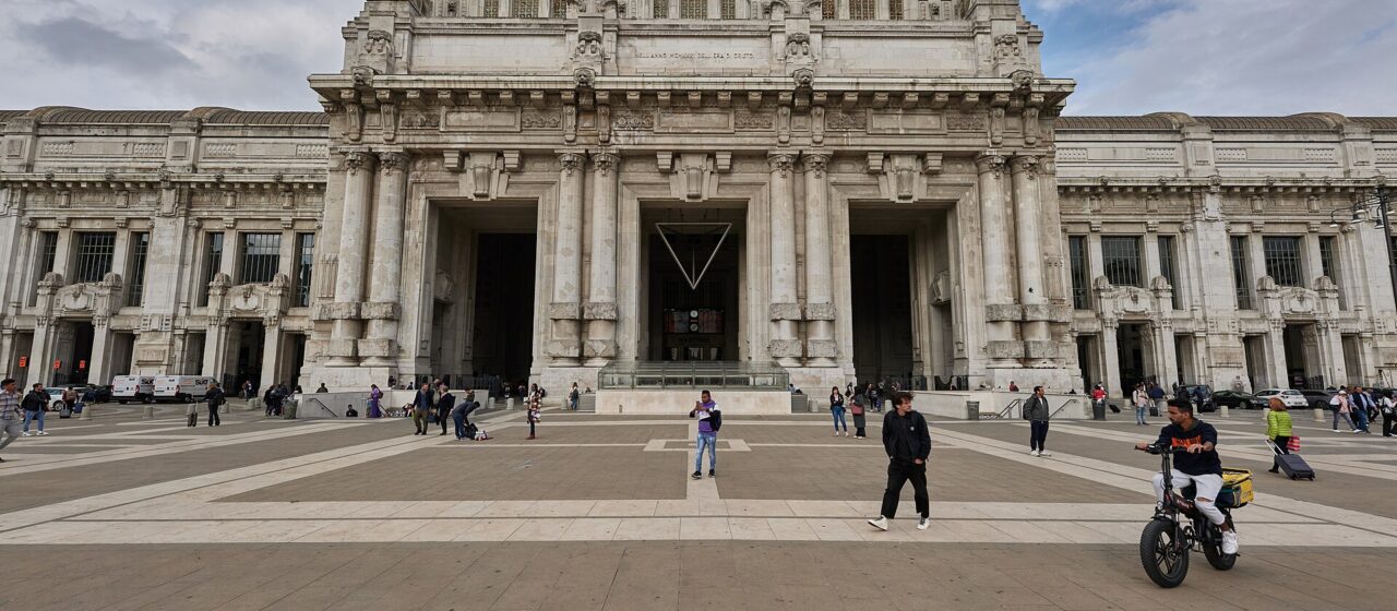 The monumental pale stone facade of Milano Centrale station seen across a wide paved square under a cloudy sky