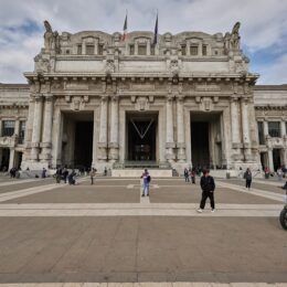 The monumental pale stone facade of Milano Centrale station seen across a wide paved square under a cloudy sky