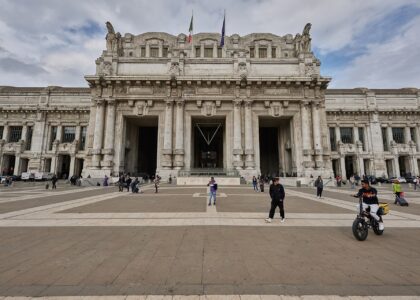 The monumental pale stone facade of Milano Centrale station seen across a wide paved square under a cloudy sky
