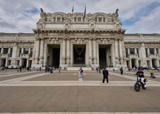 The monumental pale stone facade of Milano Centrale station seen across a wide paved square under a cloudy sky