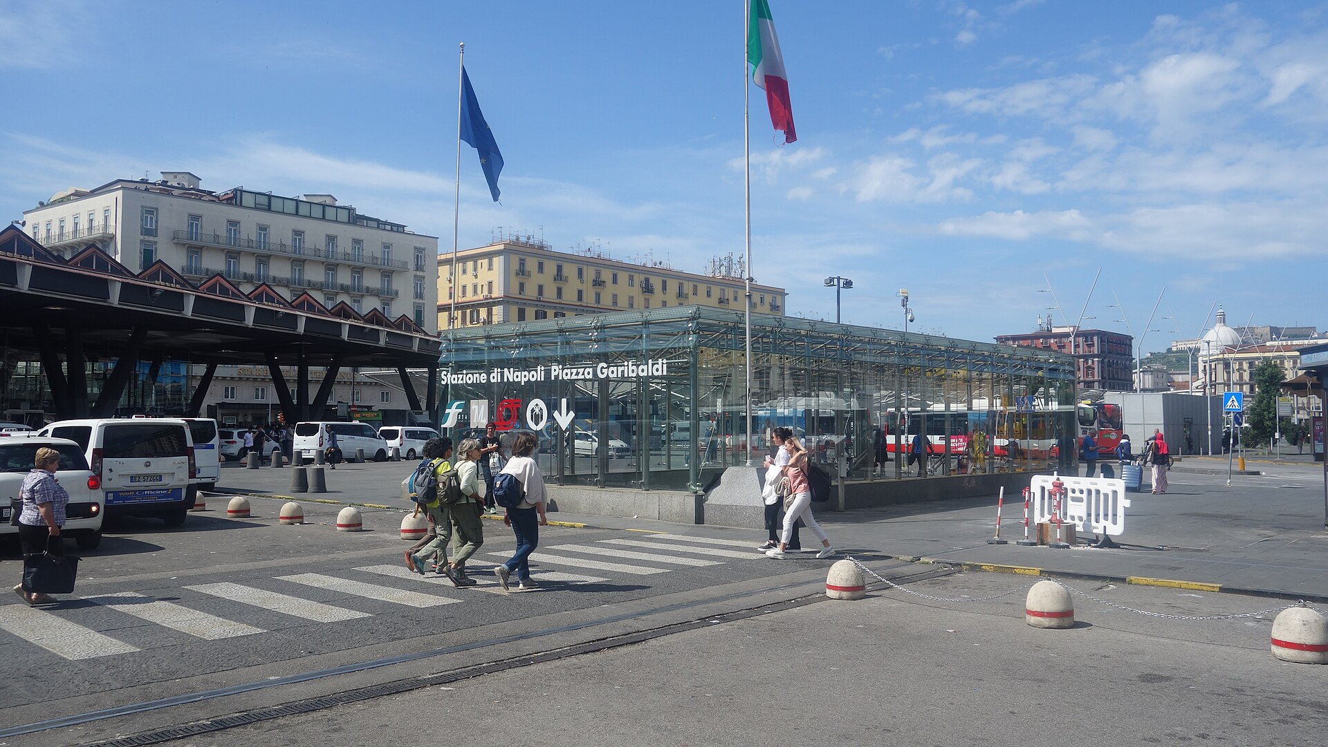 The glass entrance of Napoli Centrale station on Piazza Garibaldi, with a sign reading Stazione di Napoli Piazza Garibaldi, flags and taxis outside