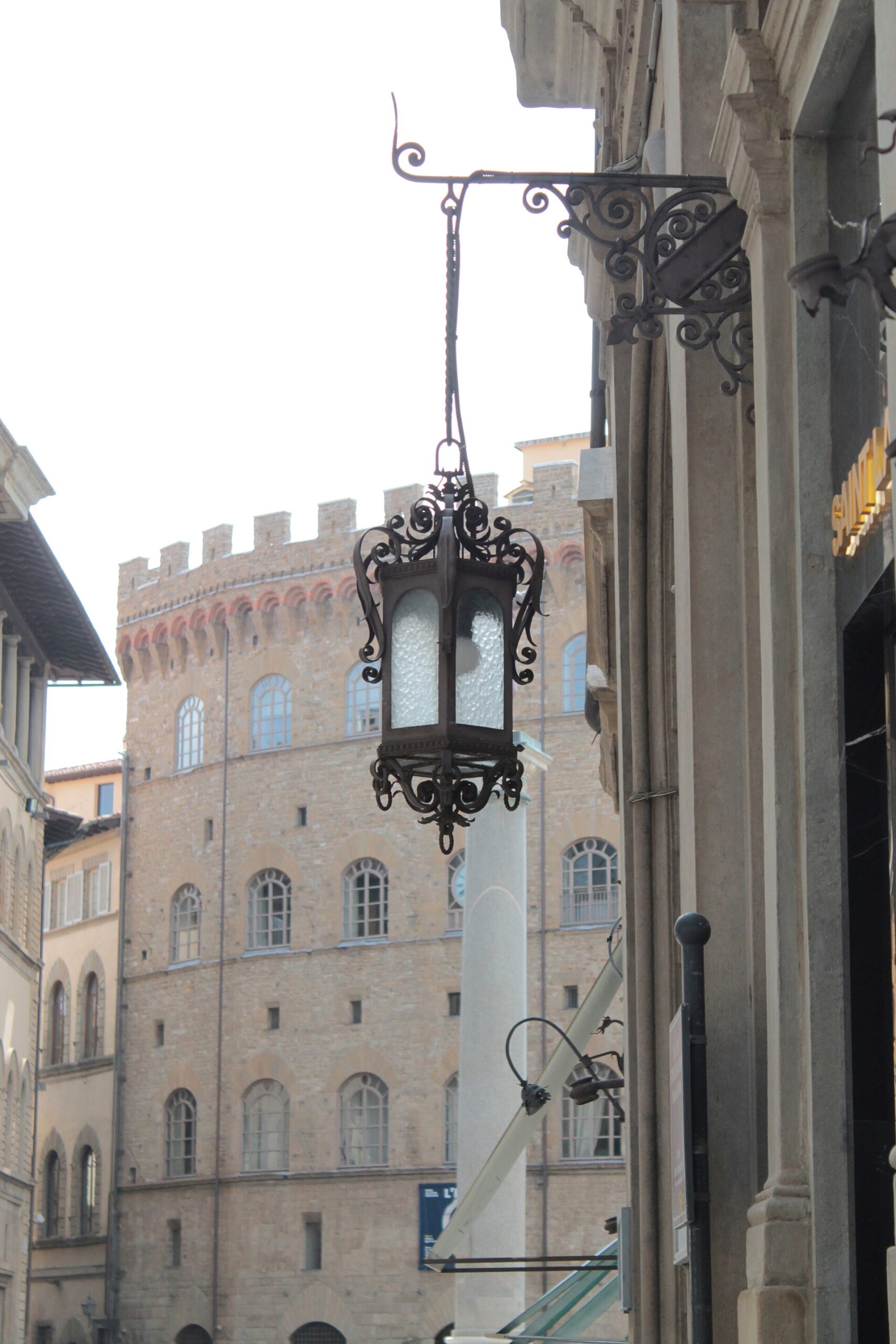 An ornate iron lantern hangs beside boutique awnings and Palazzo Spini Feroni on Florence's Via de' Tornabuoni.