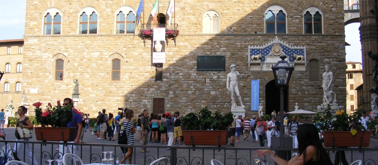 Outdoor restaurant tables with white cloths face Palazzo Vecchio and visitors in Florence's Piazza della Signoria.