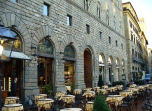 Outdoor tables with yellow cloths in front of the arched, stone-fronted Rivoire café in Florence, its name on the arched signs.