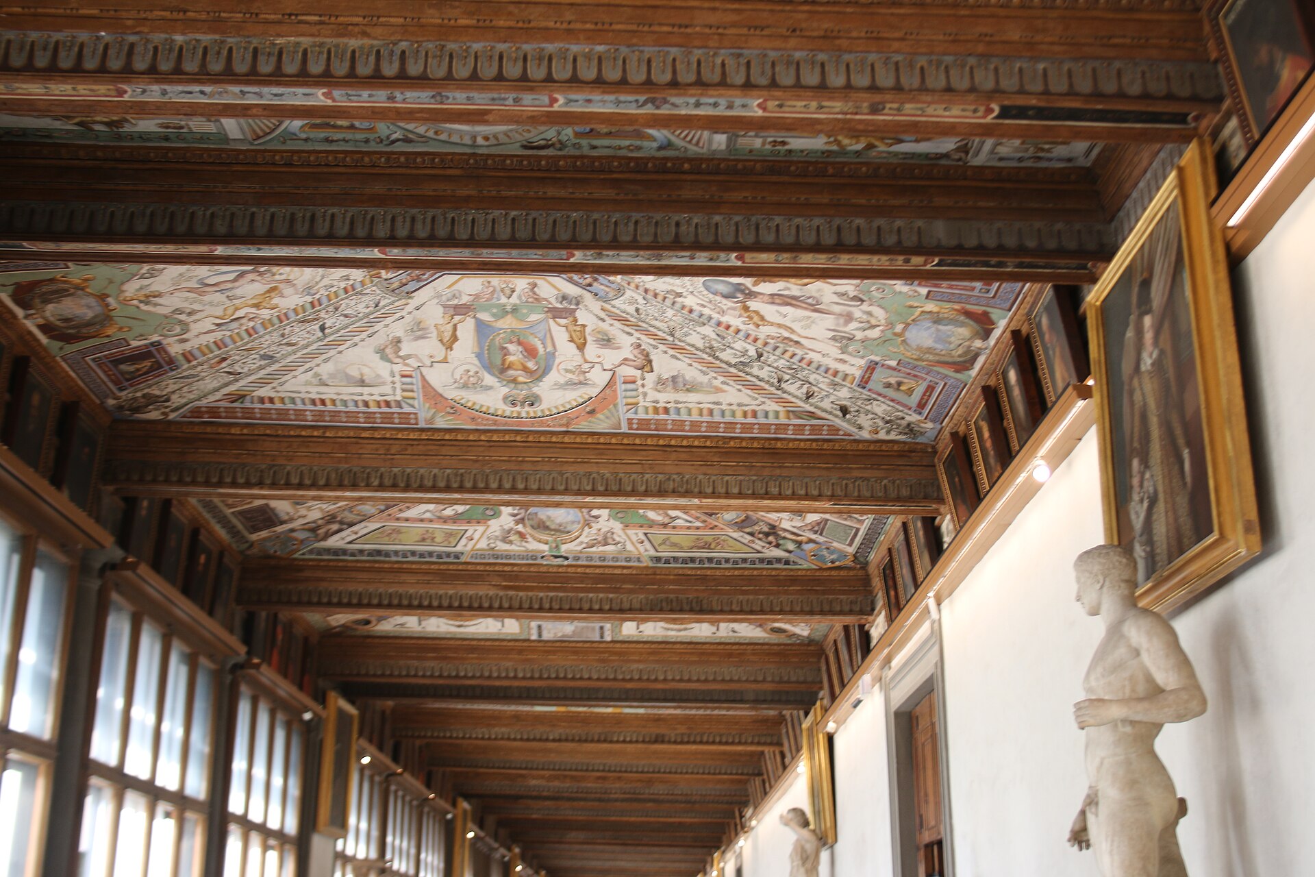 Painted ceiling panels and marble statues line a sunlit upper corridor inside the Uffizi Gallery in Florence.
