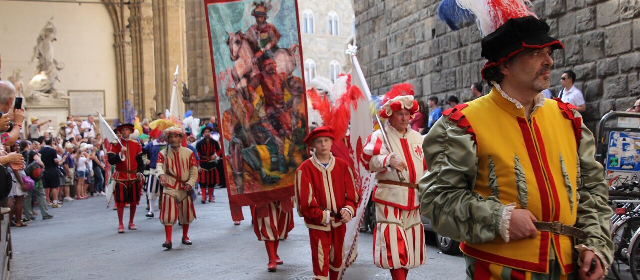 Participants in Renaissance costumes and feathered hats parade beside Florence's Loggia dei Lanzi as spectators watch.