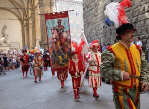 Participants in Renaissance costumes and feathered hats parade beside Florence's Loggia dei Lanzi as spectators watch.