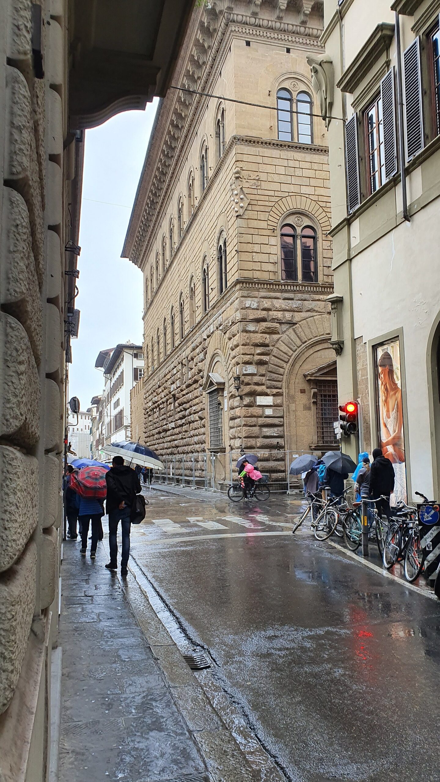 Pedestrians carry umbrellas along a rain-soaked Florence street beside Palazzo Medici Riccardi, with bicycles parked against the opposite wall.