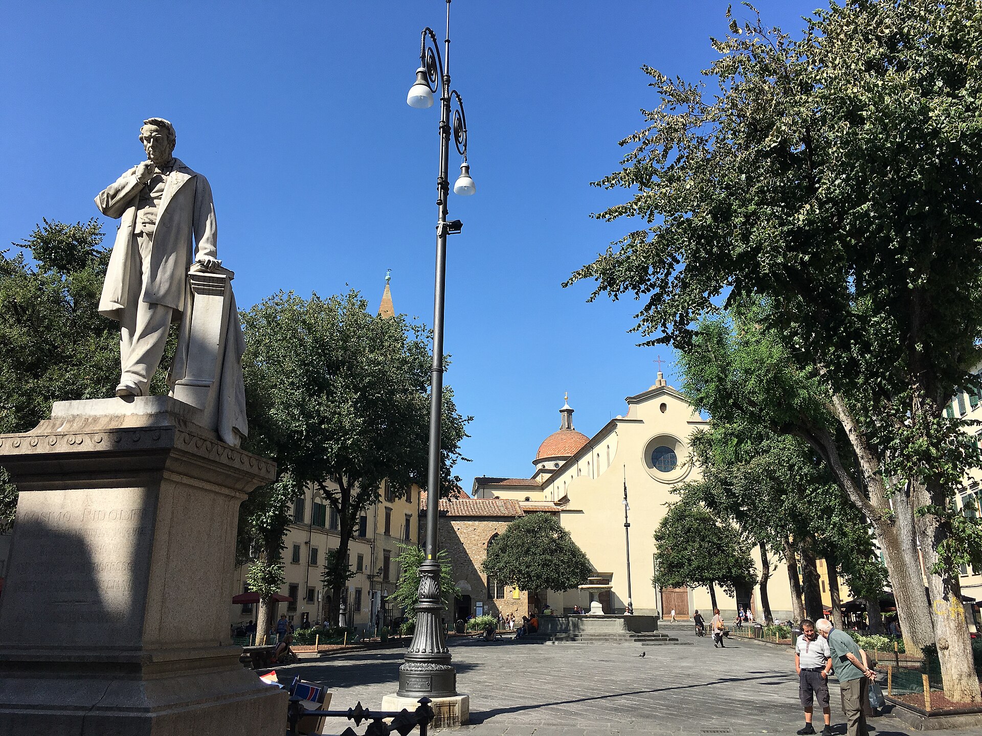 Pedestrians cross Florence’s Piazza Santo Spirito beside a statue and trees, with the church and fountain beyond.