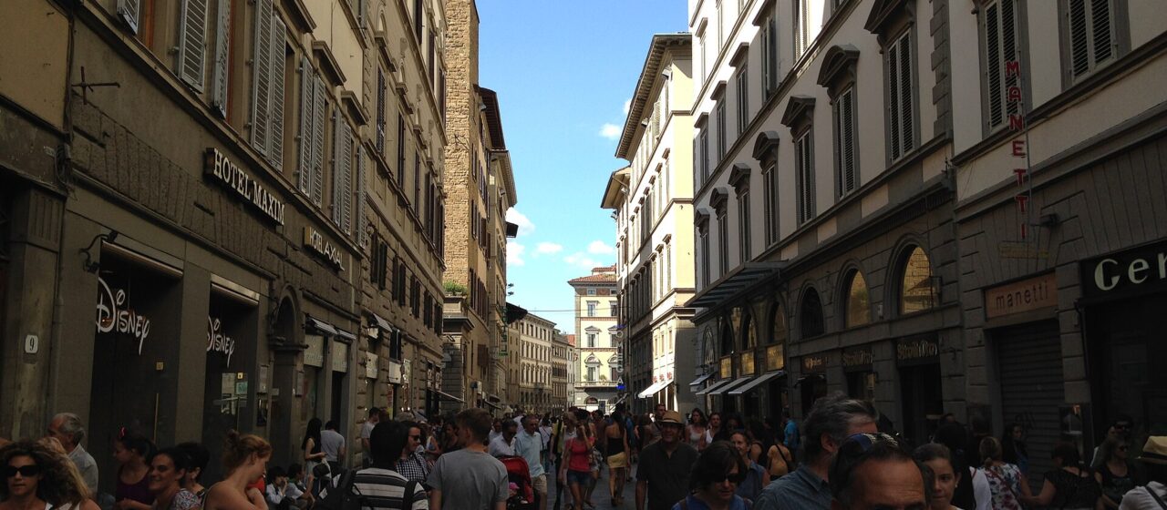 Pedestrians fill Via dei Calzaiuoli in Florence between tall shuttered buildings, with blue sky above the narrow street.