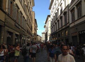 Pedestrians fill Via dei Calzaiuoli in Florence between tall shuttered buildings, with blue sky above the narrow street.