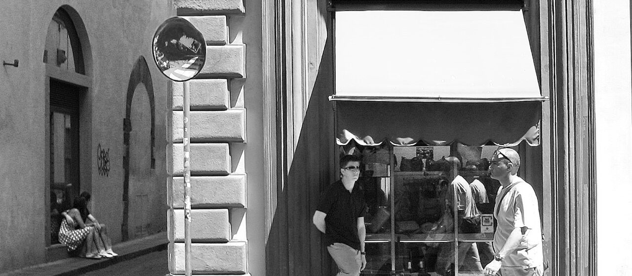 Pedestrians pass the arched Junior Leather Factory storefront in Florence in a black-and-white street scene.