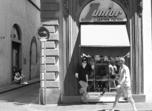 Pedestrians pass the arched Junior Leather Factory storefront in Florence in a black-and-white street scene.