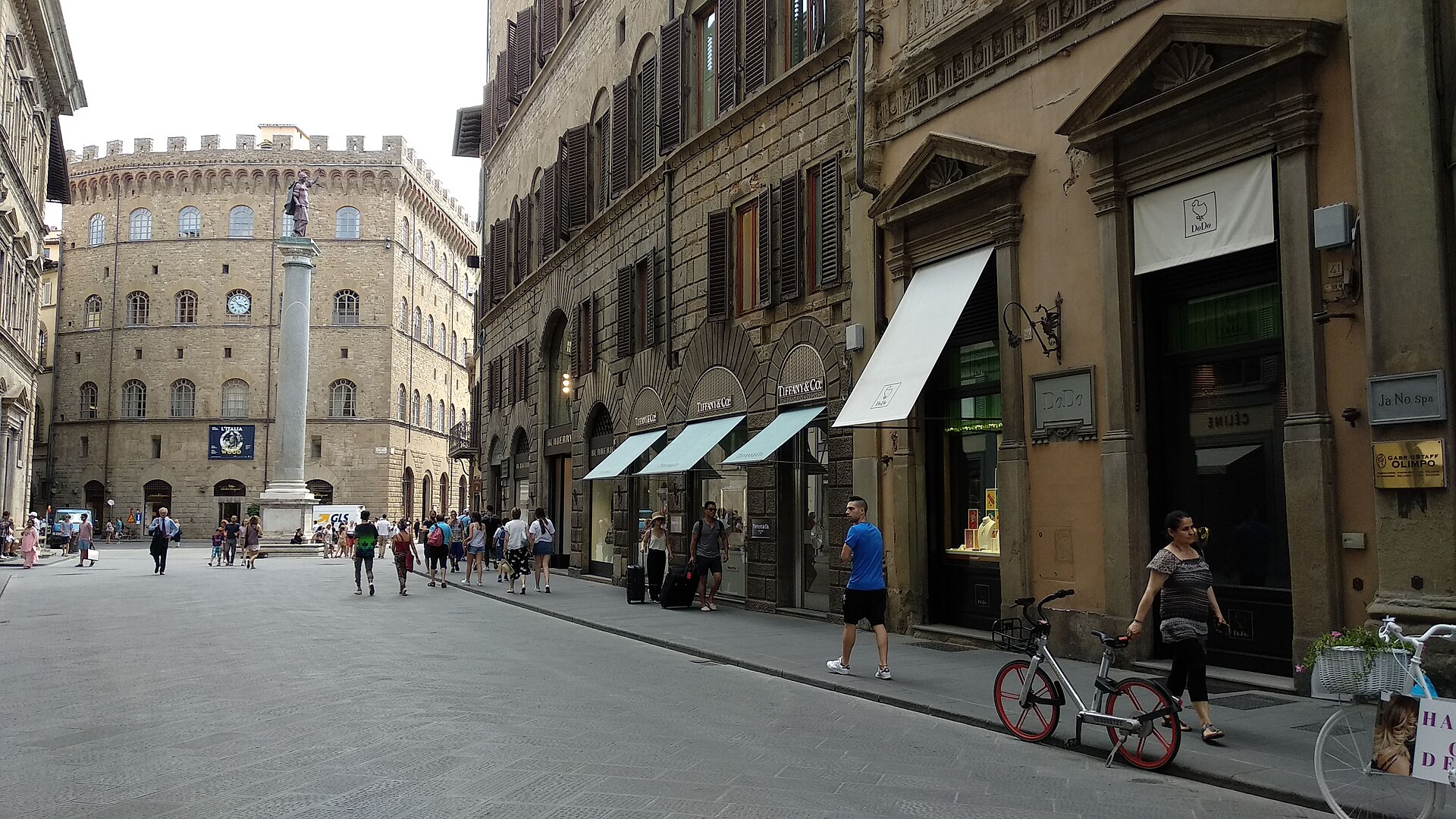 Pedestrians pass boutiques on Via de' Tornabuoni in Florence, with Palazzo Spini Feroni beyond the column.