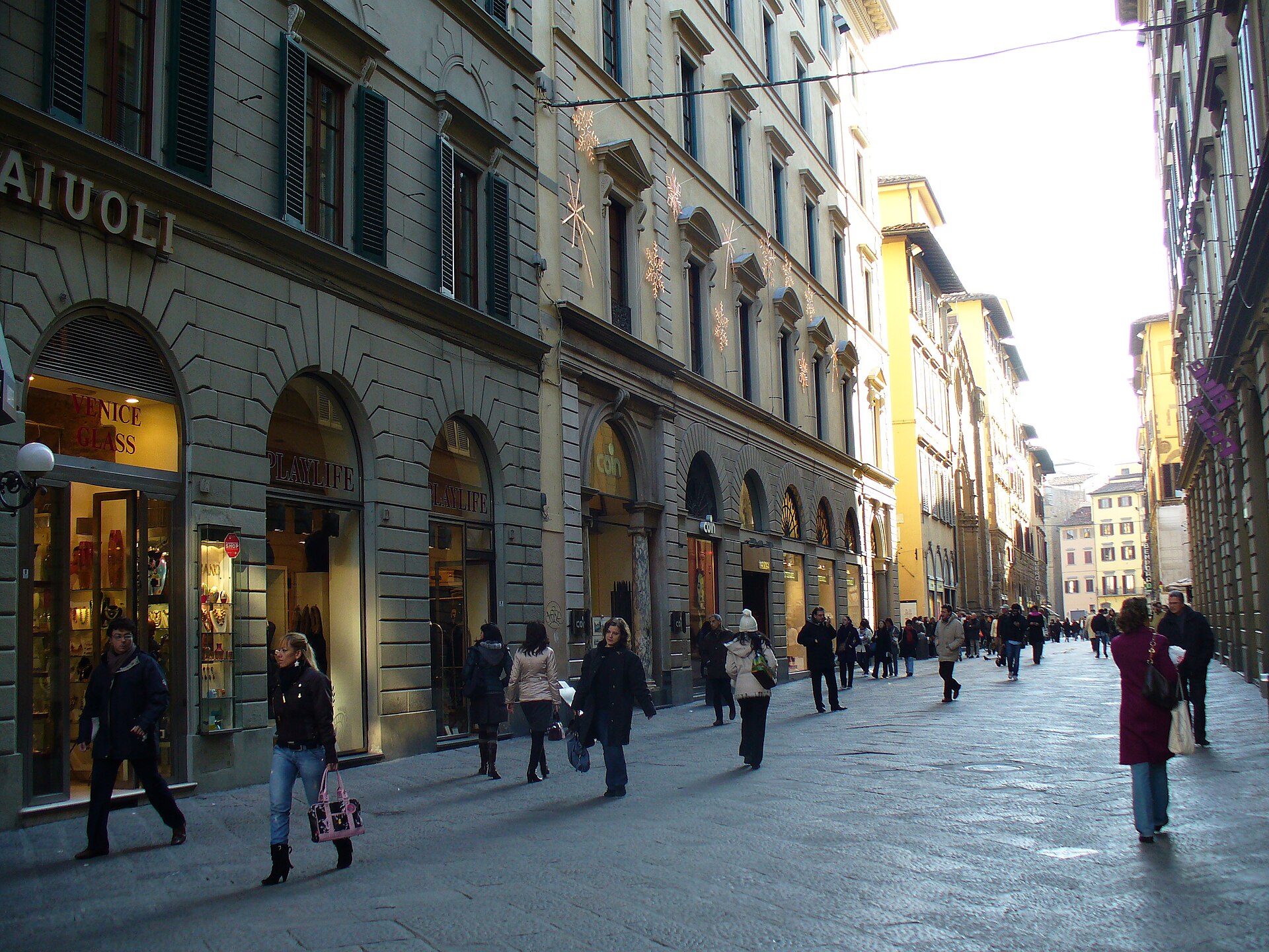 Pedestrians walk along Florence’s Via dei Calzaiuoli beneath shuttered windows and hanging star decorations, with stone paving underfoot.