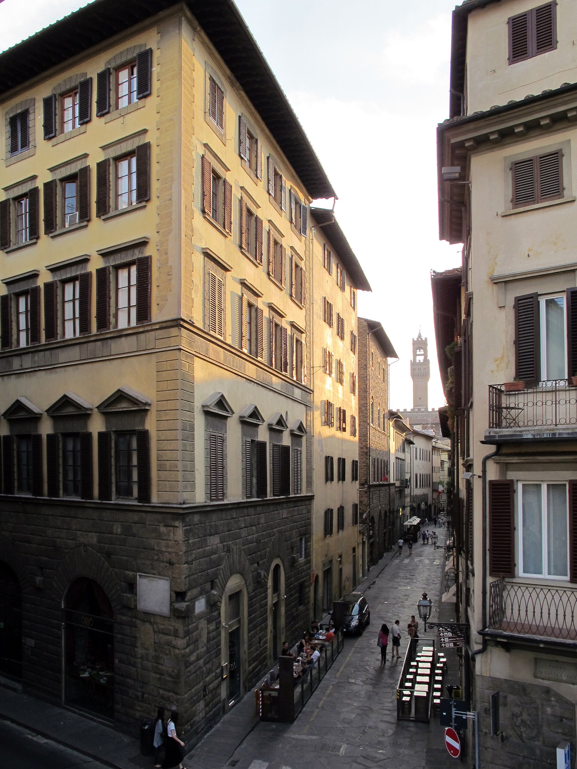 Pedestrians walk along narrow Borgo dei Greci in Florence, between shuttered buildings with Santa Croce’s bell tower beyond.