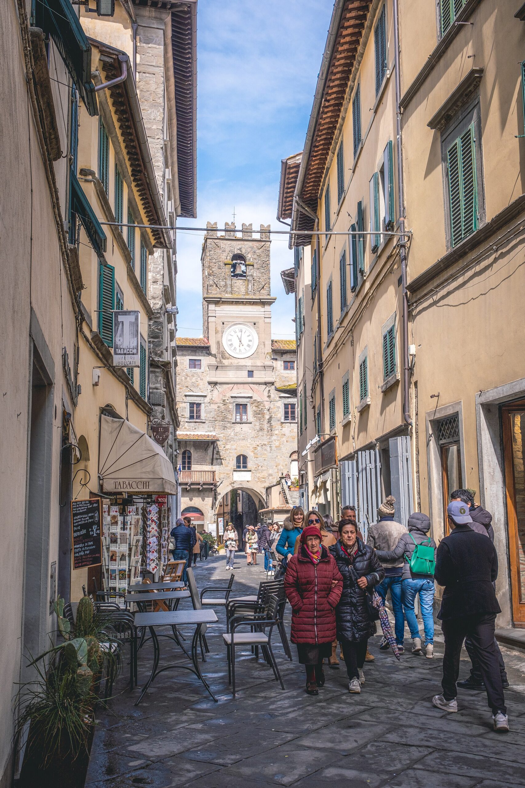Pedestrians walk along Via Nazionale in Cortona, passing outdoor tables beneath shuttered buildings with the town-hall clock tower ahead.