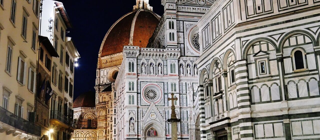 People walk beside Florence's illuminated baptistery at night, with Santa Maria del Fiore and its dome behind them.