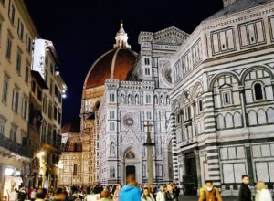 People walk beside Florence's illuminated baptistery at night, with Santa Maria del Fiore and its dome behind them.