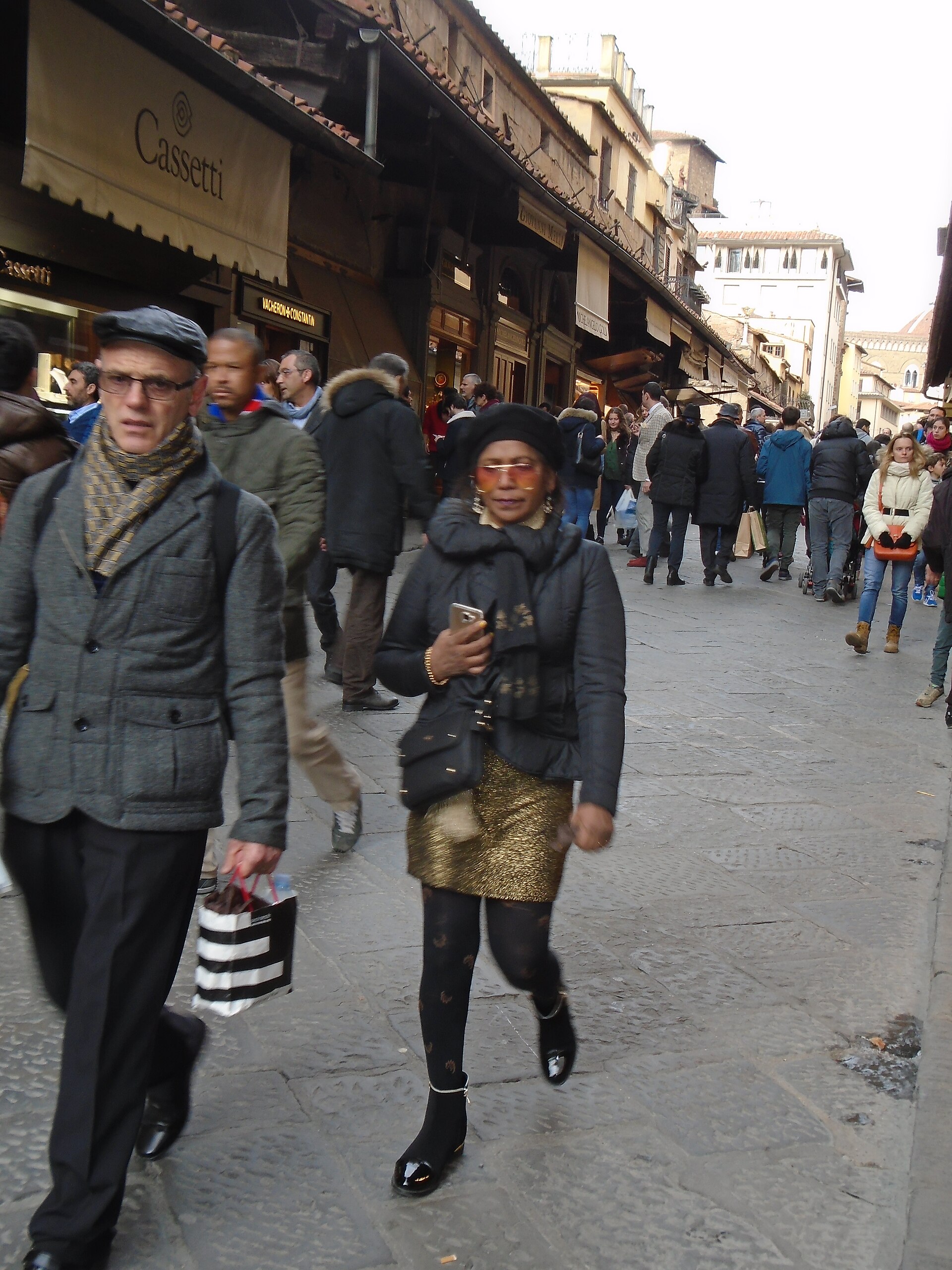 People in winter coats, hats and scarves walk past jewellery shops on Florence’s Ponte Vecchio, with more pedestrians behind them.