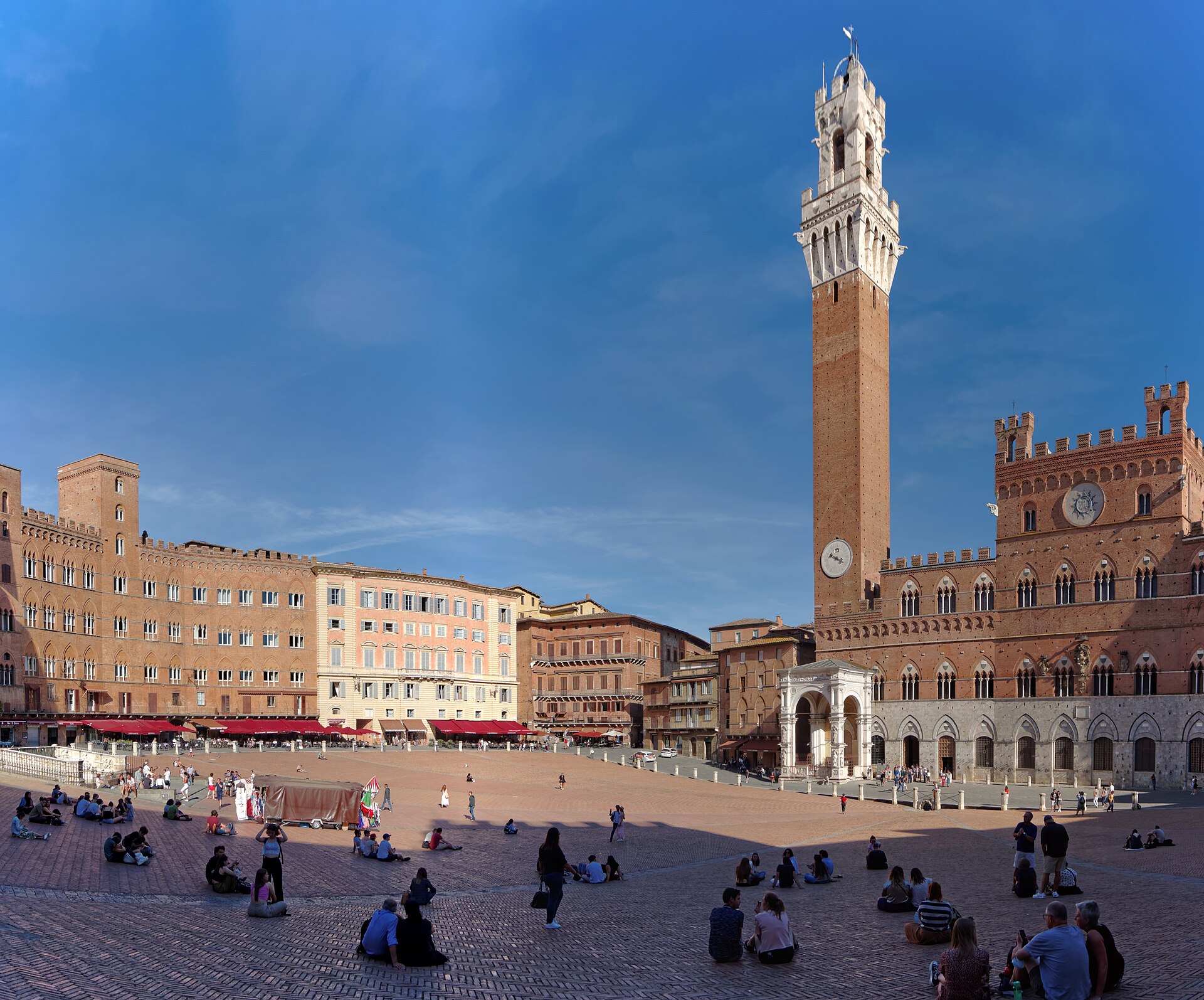 Piazza del Campo in Siena under a blue sky, with the Torre del Mangia and Palazzo Pubblico on the right and people sitting on the sloping brick paving