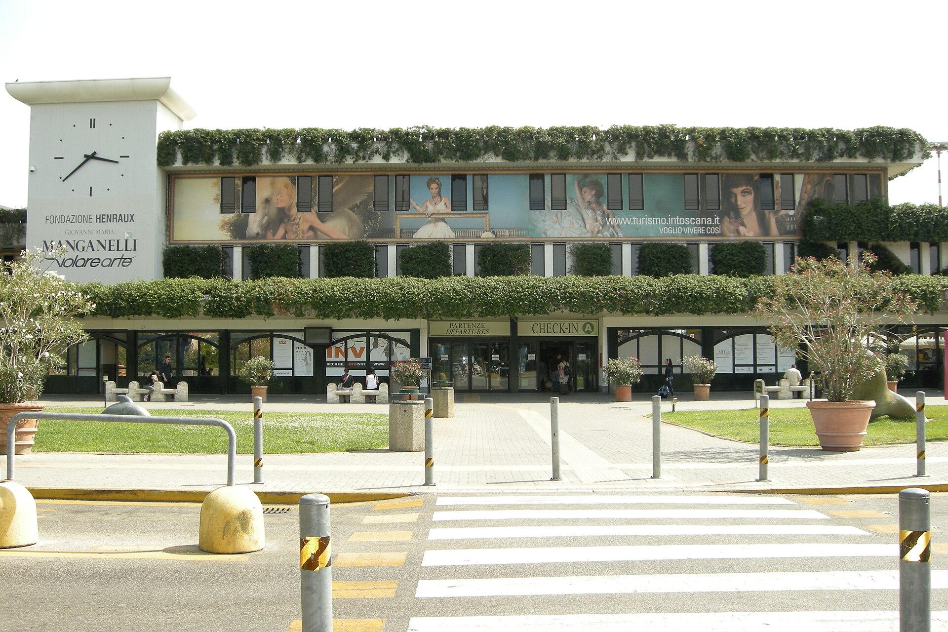 Pisa Airport’s ivy-covered terminal faces a grassy forecourt, with a clock tower and pedestrian crossing beside the entrance.