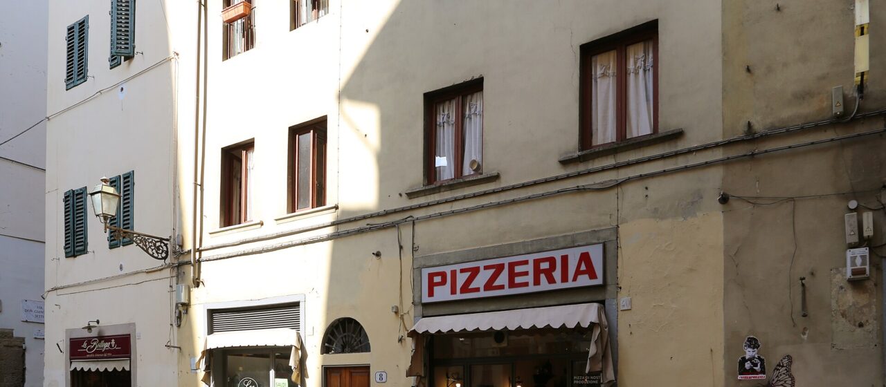 A pizzeria and neighbouring shopfronts line Via de’ Neri in Florence, with bicycles parked beneath shuttered windows.