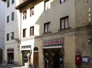 A pizzeria and neighbouring shopfronts line Via de’ Neri in Florence, with bicycles parked beneath shuttered windows.