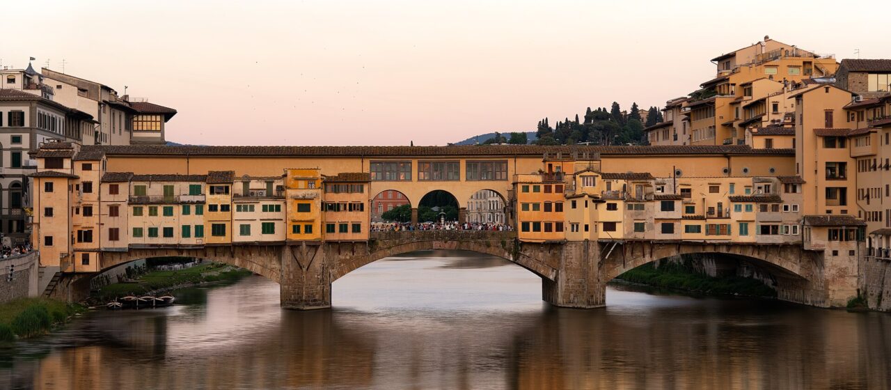 Ponte Vecchio’s projecting shops and three stone arches reflect in the Arno beneath a pale pink evening sky in Florence.