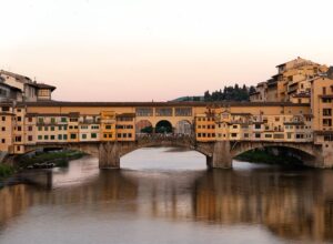 Ponte Vecchio’s projecting shops and three stone arches reflect in the Arno beneath a pale pink evening sky in Florence.