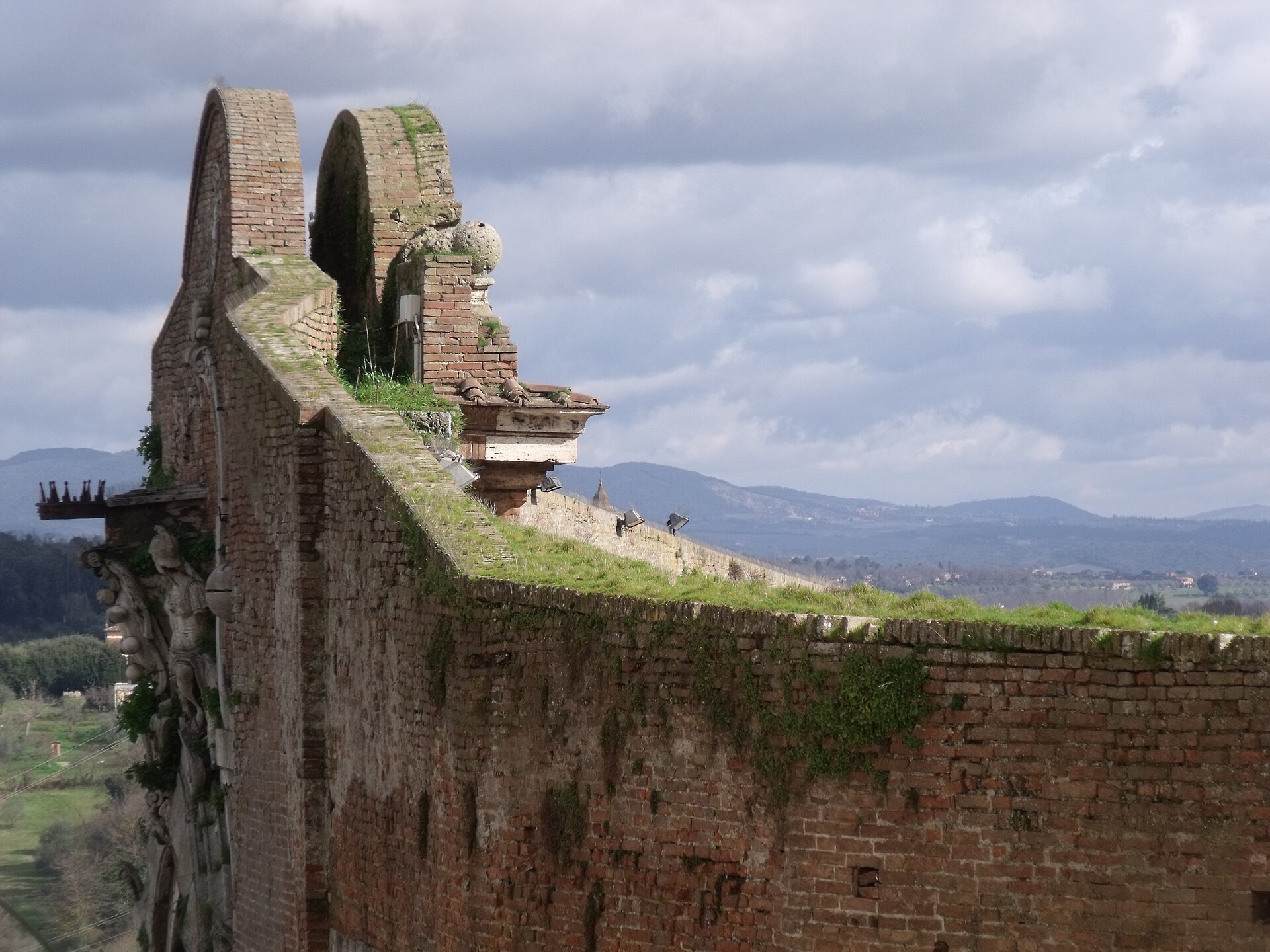 The brick top of Porta Camollia, Siena's northern gate, with grass growing on the walls and green Tuscan hills under a cloudy sky beyond