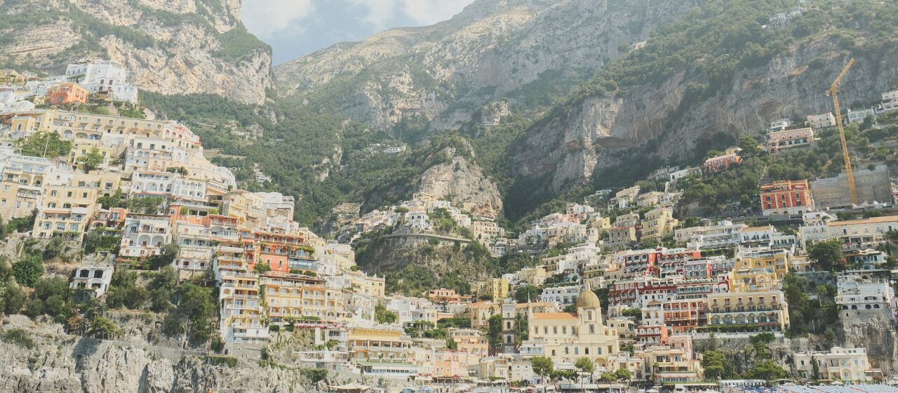 Positano seen from the sea, its pastel houses stacked up a steep green hillside above a small bay with boats moored in the water