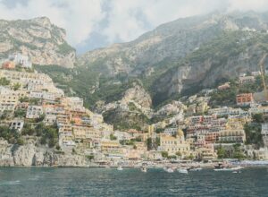 Positano seen from the sea, its pastel houses stacked up a steep green hillside above a small bay with boats moored in the water