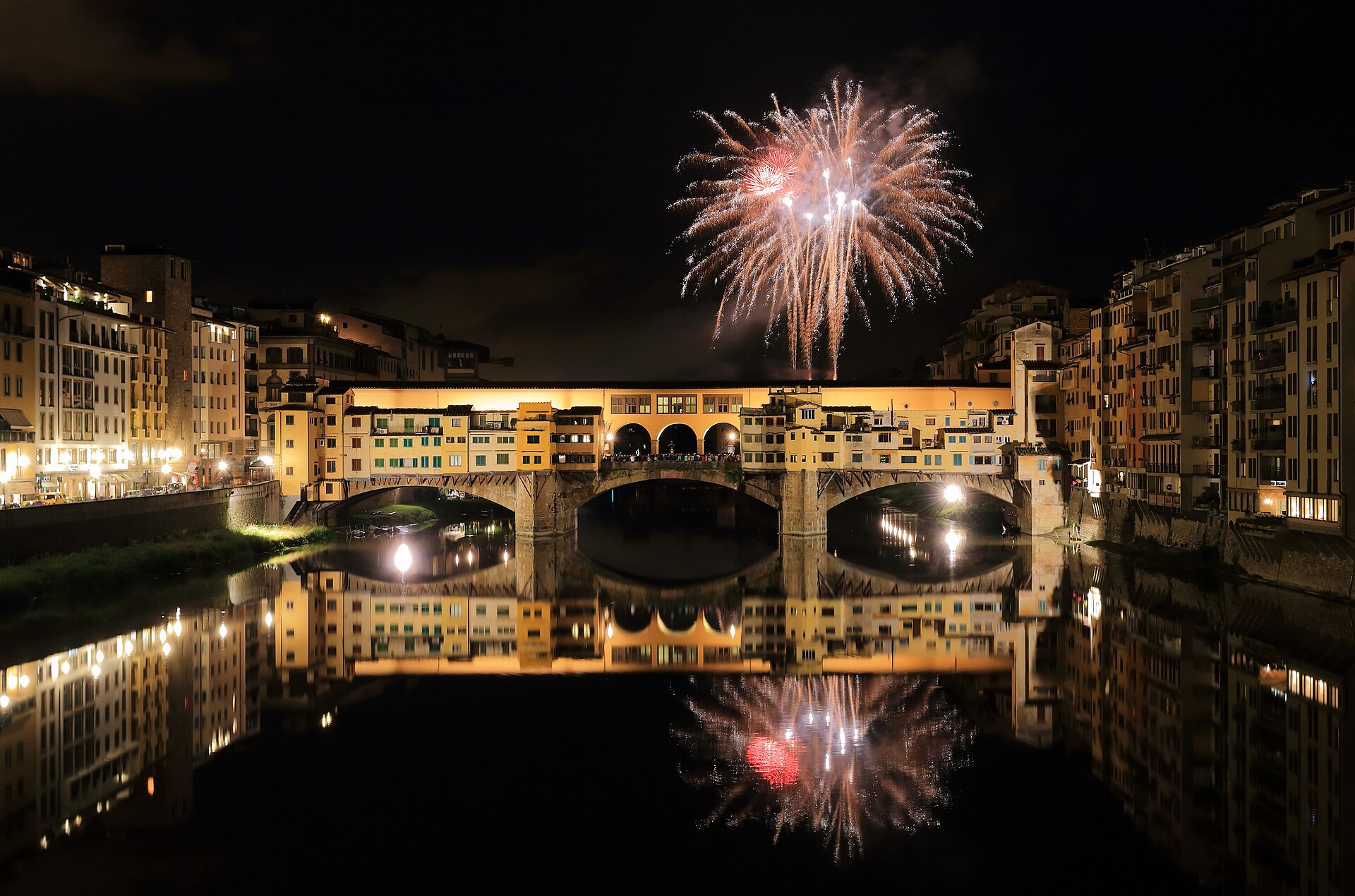 Red and white fireworks burst over Florence's illuminated Ponte Vecchio and reflect in the dark water of the Arno.