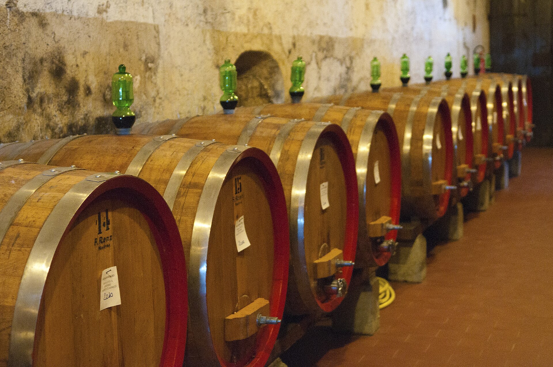 Rows of wooden wine barrels line a stone-walled cellar at Vignamaggio in Chianti, with green airlocks above them.