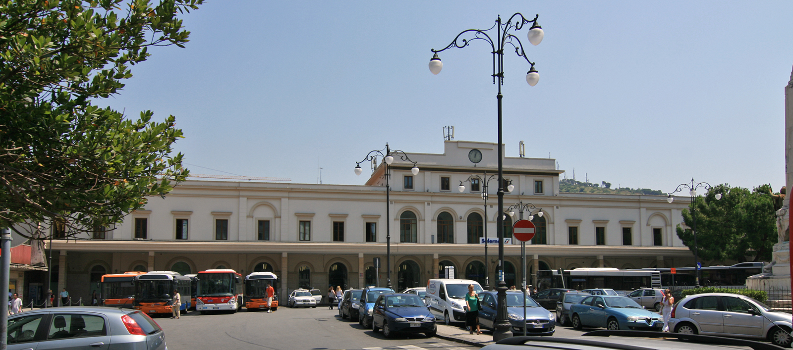The pale arcaded facade of Salerno railway station with orange buses and parked cars in the forecourt and an ornate lamp post in front