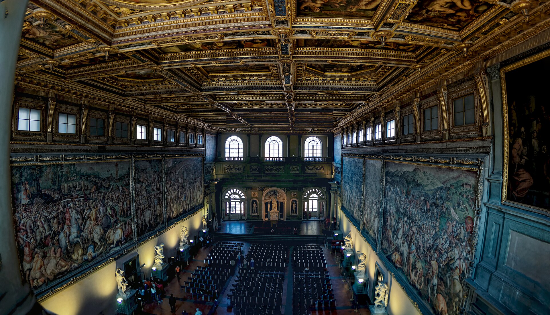 The Salone dei Cinquecento in Florence's Palazzo Vecchio displays vast battle frescoes beneath a gilded coffered ceiling above rows of chairs.